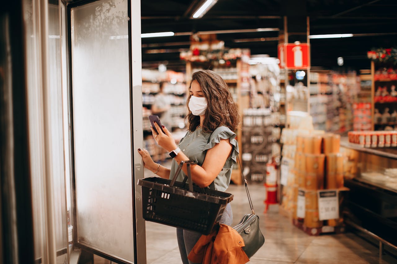 Woman wearing mask shopping with a basket and smartphone in a supermarket.